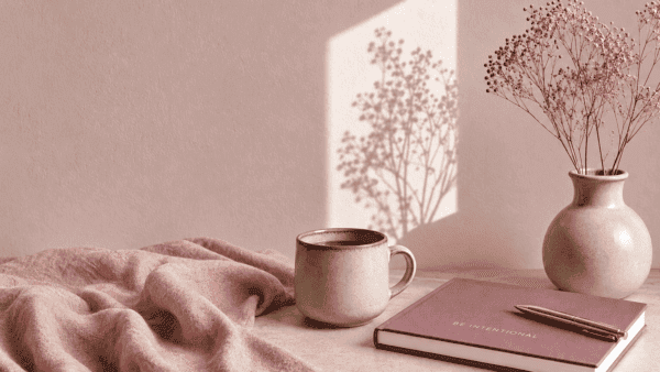 Warm pastel still life with a ceramic mug, notebook, pen, dried flowers, and soft sunlight on a pink wall.